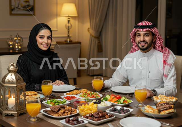 مائده طعام مليئه بأصناف المأكولات و المقبلات الشهية، ديكور وأجواء رمضانية، النظر الى الكاميرا بايماءات البهجة والسرور، رجل عربي خليجي سعودي يرتدي الشماغ والثوب التقليدي يجلس بجانب زوجته حول سفرة الإفطار في شهر رمضان المبارك، الألفة والمحبة بين الزوجين