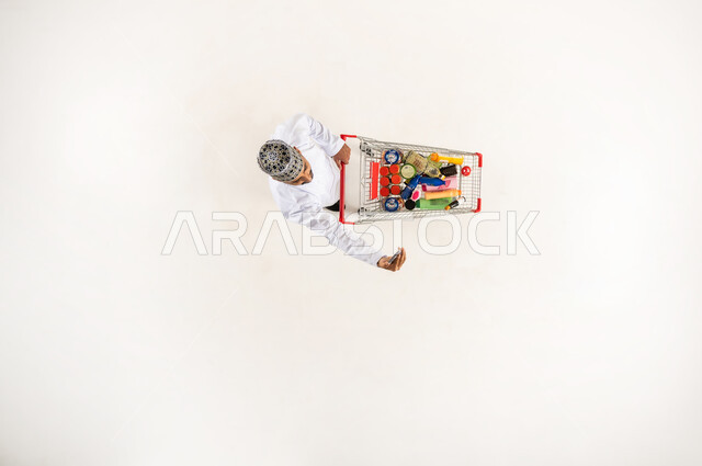 استخدام جهاز تقني حديث متطور, شراء المنتجات و المستلزمات المنزلية التموينيه من المراكز التجارية, بورتريه عامودي علوي لرجل عربي خليجي عماني يرتدي الكمه و الدشداشة يلتقط صور السيلفي عبر الهاتف المحمول يمسك بيده عربة التسوق في السوبر ماركت, خلفيه بيضاء