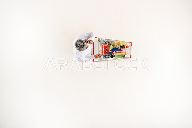 السؤال عن المنتجات اللازمة المنزلية التموينيه من المراكز التجارية, الاستفادة من الخصومات و التخفيضات والعروض, بورتريه عامودي علوي لرجل عربي خليجي عماني يرتدي الكمه و الدشداشة يقوم باجراء مكالمة هاتفيه عبر الهاتف المحمول يجر عربة التسوق في السوبر ماركت, خلفيه بيضاء