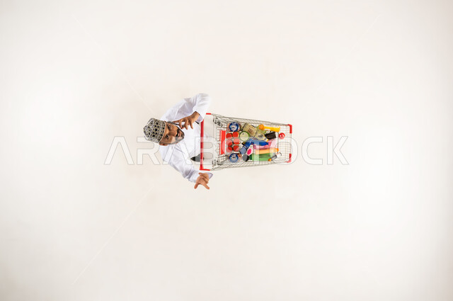 النظر الى الاعلى بايماءات السؤال عن المنتجات التموينية, الاستفادة من التخفيضات و الخصومات, بورتريه عامودي علوي لرجل عربي خليجي عماني يرتدي الكمه و الدشداشة يقوم باجراء مكالمة هاتفيه عبر الهاتف المحمول يجر عربة التسوق في السوبر ماركت, خلفيه بيضاء