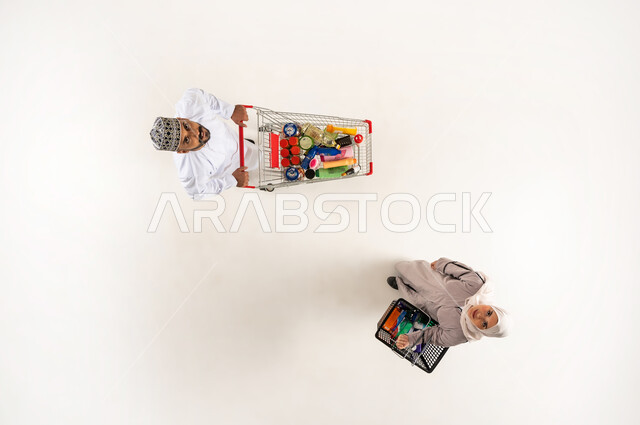 الاستفادة من الخصومات و التخفيضات والعروض على المنتجات, شاب يقوم بجر عربة التسوق, البحث عن المستلزمات المنزلية التموينيه من المراكز التجارية العمانية, بورتريه عامودي علوي لرجل و امرأة  عربيان خليجيان عمانيان يقومان بالتسوق و الشراء من السوبر ماركت, خلفيه بيضاء