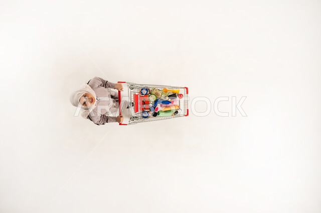 Saudi woman taking advantage of discounts and reductions on products, spending free time buying household supplies from shopping malls, vertical top portrait of a veiled Gulf Omani Arab woman wearing a beige abaya pulling a shopping cart in a supermarket, white background