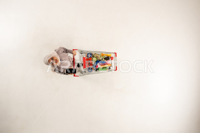 سيدة سعودية تستخدم جهاز تقني حديث متطور, شراء المنتجات و المستلزمات المنزلية من المراكز التجارية, بورتريه عامودي علوي لامرأة عربية خليجية عمانية محجبة ترتدي العباءه البيج تقوم بعرض شاشه سوداء فارغه عبر الجوال تمسك بيدها عربة التسوق في السوبر ماركت, خلفيه بيضاء