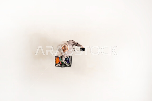 Taking advantage of discounts and reductions on products, a Saudi woman buying household supplies from shopping malls, vertical portrait of a veiled Gulf Omani Arab woman wearing a beige abaya holding a shopping basket and a mobile phone in the supermarket, white background