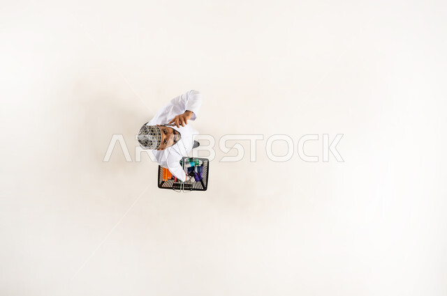 اجراء مكالمة هاتفيه عبر الجوال, استعمال جهاز تقني حديث, شراء المستلزمات المنزلية التموينيه من المراكز التجارية, الاستفادة من الخصومات و التخفيضات, بورتريه عامودي علوي لشاب عربي خليجي عماني يرتدي الكمه و الدشداشة يحمل بيده سلة التسوق في السوبر ماركت, خلفيه بيضاء
