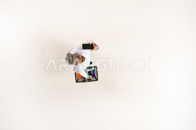 عرض شاشه سوداء فارغه عبر الجوال, استعمال جهاز تقني حديث, شراء المستلزمات المنزلية التموينيه من المراكز التجارية, الاستفادة من الخصومات و التخفيضات, بورتريه عامودي علوي لشاب عربي خليجي عماني يرتدي الكمه و الدشداشة يحمل بيده سلة التسوق في السوبر ماركت, خلفيه بيضاء