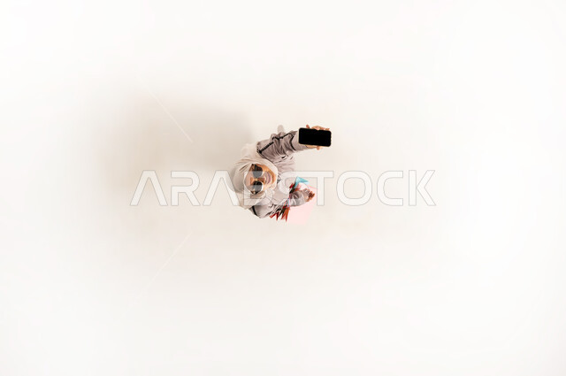Blank black screen view on mobile phone, using modern advanced technical device, vertical overhead portrait of veiled Omani Gulf young woman wearing beige abaya and sunglasses holding colorful shopping bags, Saudi lady looking at camera with happy expression, white background