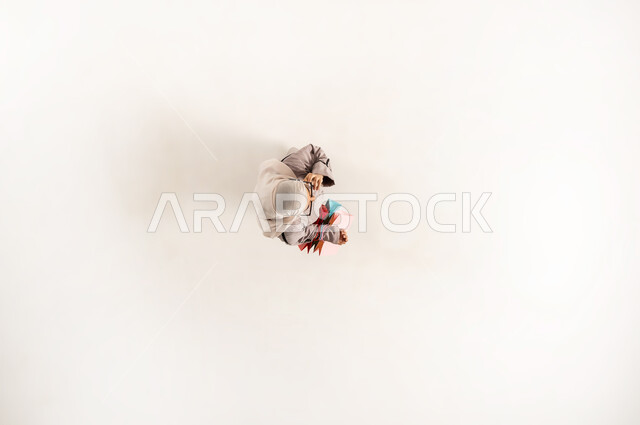 Saudi woman making a phone call via mobile phone, shopping from shops and malls, communicating with family and friends, vertical overhead portrait of a veiled young Omani Gulf woman wearing a beige abaya carrying colorful shopping bags, using a modern advanced technical device, white background