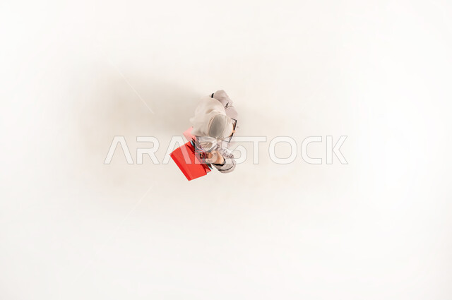 الاستمتاع في الشراء من المراكز التجاريه, عروض وتخفيضات موسمية في المولات، بورتريه علوي عامودي لإمرأة عربية خليجية عمانية محجبة ترتدي العباءة تحمل بيدها أكياس تسوق ملونة، سيدة سعودية تقوم بالترويج للمنتجات العمانية والماركات العالمية الفاخرة، خلفية بيضاء