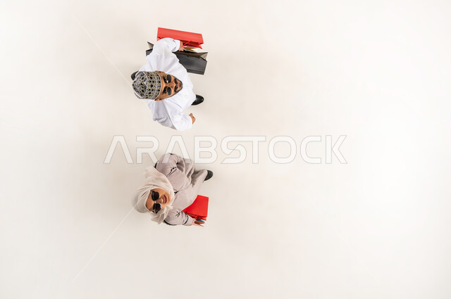 النظر الى الكاميرا بإيماءات السرور، قضاء وقت ممتع بالتسوق، الاستفادة من الحسم والتخفيض، بورتريه علوي عامودي لشاب عربي خليجي عماني يرتدي الدشداشة والكمه يقف مع زوجته يرتديان النظارة الشمسية يمسكان أكياس ورقية ملونة، سيدة محجبة ترتدي العباءة، خلفية بيضاء