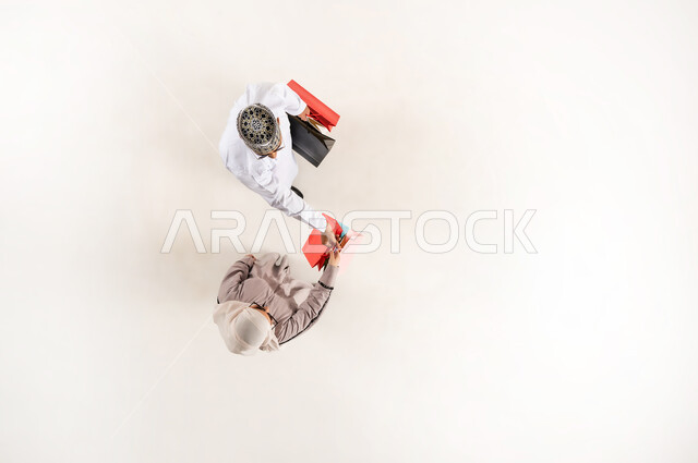 الاستفادة من الحسم والتخفيض، قضاء وقت ممتع بالتسوق، بورتريه علوي عامودي لشاب عربي خليجي عماني يرتدي الدشداشة والكمه والنظارة الشمسية يمسك بيده أكياس ورقية ملونة يقدم الهدايا لسيدة محجبة ترتدي العباءة، الشعور بالفرح والسرور، خلفية بيضاء