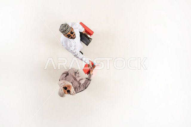 قضاء وقت ممتع بالتسوق، الاستفادة من الحسم والتخفيض، بورتريه علوي عامودي لشاب عربي خليجي عماني يرتدي الدشداشة والكمه والنظارة الشمسية يمسك بيده أكياس ورقية ملونة يقدم الهدايا لسيدة محجبة ترتدي العباءة، النظر الى الكاميرا بإيماءات السرور، خلفية بيضاء