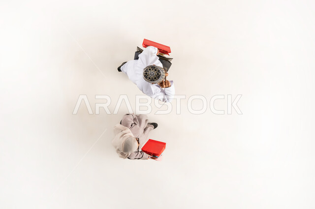 Online shopping concept, use of technology and advanced technology, vertical overhead portrait of a young Arab Gulf Omani man wearing a dishdasha, a sleeve and sunglasses making a voice call via mobile, a veiled woman holding colorful paper bags in her hand, white background