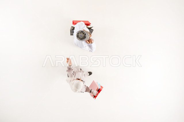 Taking advantage of the discount and reduction, having a good time shopping, vertical overhead portrait of a young Arab Gulf Omani man wearing a dishdasha, a sleeve and sunglasses, putting his hand on his head with thinking gestures, a veiled woman holding colorful paper bags, white background