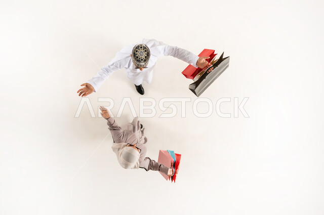 الشعور بالفرح والحماس للاستفادة من الحسم والتخفيض، قضاء وقت ممتع بالتسوق، بورتريه علوي عامودي لشاب عربي خليجي عماني يرتدي الدشداشة والكمه والنظارة الشمسية مع سيدة محجبة يحملان أكياس ورقية ملونة، خلفية بيضاء