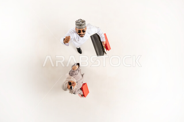 Taking advantage of the discount and reduction, having a good time shopping, vertical overhead portrait of a young Arab Gulf Omani man wearing a dishdasha, a turban and sunglasses with a veiled woman holding colorful paper bags looking at the camera with gestures of pleasure, white background
