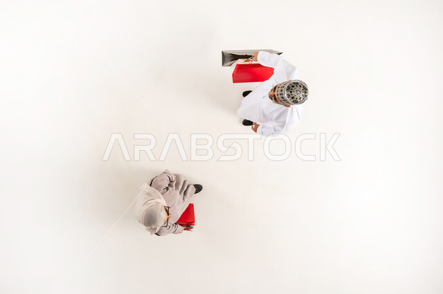 قضاء وقت ممتع بالتسوق، الاستفادة من الحسم والتخفيض، بورتريه علوي عامودي لشاب عربي خليجي عماني يرتدي الدشداشة والكمه والنظارة الشمسية مع سيدة محجبة يحملان أكياس ورقية ملونة ينظران الى الكاميرا بإيماءات السرور، خلفية بيضاء