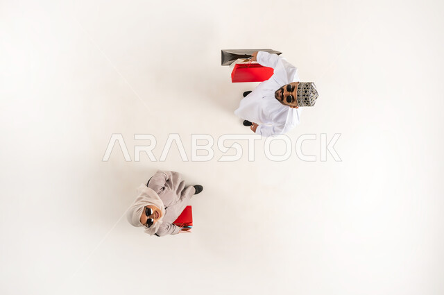 الاستفادة من الحسم والتخفيض، قضاء وقت ممتع بالتسوق، بورتريه علوي عامودي لشاب عربي خليجي عماني يرتدي الدشداشة والكمه والنظارة الشمسية مع سيدة محجبة يحملان أكياس ورقية ملونة ينظران الى الكاميرا بإيماءات السرور، خلفية بيضاء