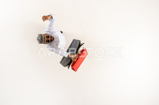 الاستفادة من الحسم والتخفيض، بورتريه علوي عامودي لشاب عربي خليجي عماني يرتدي الدشداشة والكمه والنظارة الشمسية يمسك بيده أكياس ورقية ملونة يرفع يده للاعلى بتعابير الفرح، النظر الى الكاميرا بإيماءات السرور، قضاء وقت ممتع بالتسوق، خلفية بيضاء