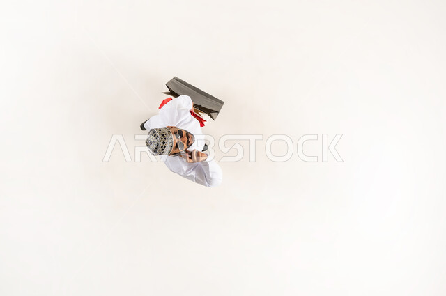 Completing the order process using a mobile phone, the concept of online shopping and purchasing, vertical overhead portrait of a young Arab Gulf Omani man wearing a dishdasha, a sleeve and sunglasses holding colorful paper bags in his hand making a voice call via mobile phone, white background