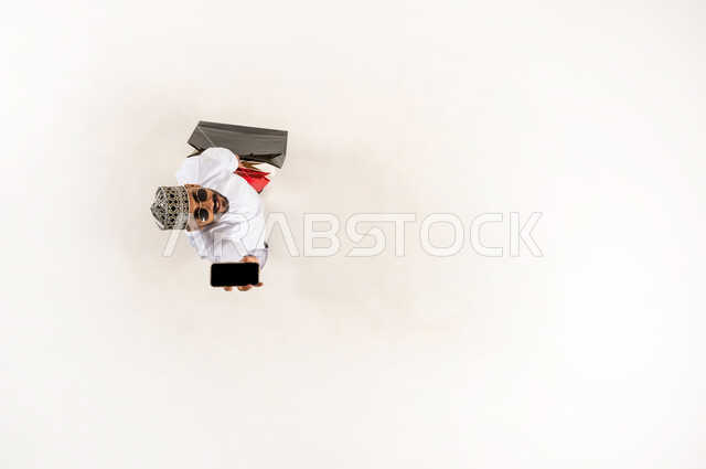 Online shopping concept, completing order using mobile phone, vertical overhead portrait of young Arab Gulf Omani man wearing dishdasha, sleeve and sunglasses holding colorful paper bags displaying blank black screen via mobile phone, looking at camera with gestures of pleasure, white background