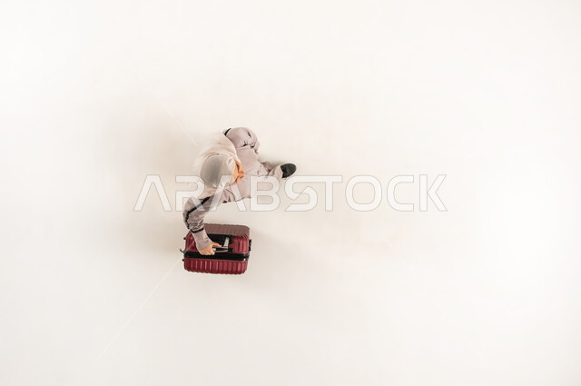 قضاء وقت ممتع في الإجازة, مفهوم السياحة والسفر, الاستعداد لموعد الرحلة، بورتريه علوي عامودي لشابة عربية خليجية عمانية محجبة ترتدي العباءة تمسك بيدها حقيبة السفر، سيدة سعودية تشعر بالحماس والسرور، خلفية بيضاء