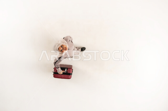 سيدة سعودية تنظر الى الكاميرا بإيماءات السرور، قضاء وقت ممتع في الإجازة, مفهوم السياحة والسفر, الاستعداد لموعد الرحلة، بورتريه علوي عامودي لشابة عربية خليجية عمانية محجبة ترتدي العباءة تمسك بيدها حقيبة السفر، الشعور بالحماس والفرح، خلفية بيضاء