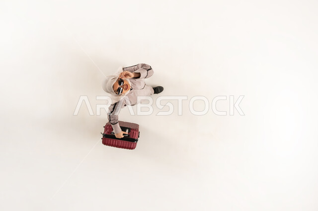 Using modern technology and tourism application, Saudi woman looking at camera with gestures of pleasure, vertical overhead portrait of veiled young Arab Gulf Omani woman wearing abaya and sunglasses holding suitcase making voice call on mobile phone, white background