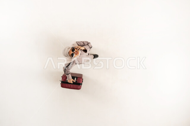 استخدام التقنية الحديثة والتطبيق السياحي, سيدة سعودية تنظر الى الكاميرا بإيماءات السرور, بورتريه علوي عامودي لشابة عربية خليجية عمانية مبتسمة محجبة ترتدي العباءة تمسك حقيبة السفر تقوم بإجراء مكالمة صوتية عبر الهاتف المحمول, خلفية بيضاء