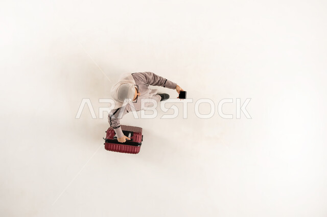 إجراء مكالمة فيديو، الخدمة المتطورة لبرنامج السياحة والسفر, بورتريه علوي عامودي لشابة عربية خليجية عمانية محجبة ترتدي العباءة و النظارة الشمسية تمسك حقيبة السفر و الهاتف المحمول, سيدة سعودية تقوم بالتقاط صورة السيلفي التذكارية، خلفية بيضاء