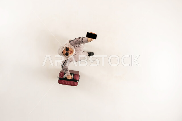 سيدة سعودية تنظر الى الكاميرا بإيماءات السرور, الخدمة المتطورة لبرنامج السياحة والسفر, بورتريه علوي عامودي لشابة عربية خليجية عمانية مبتسمة محجبة ترتدي العباءة و النظارة الشمسية تمسك حقيبة السفر تقوم بعرض الهاتف المحمول بشاشة سوداء فارغة, خلفية بيضاء