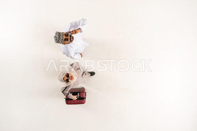 الاستعداد لرحلة ترفيهية ممتعة, النظر الى الكاميرا بإيماءات السرور، الخدمة المتطورة لبرنامج السياحة والسفر, بورتريه علوي عامودي لشاب عربي خليجي عماني يرتدي الدشداشة و الكمه يقوم بإجراء مكالمة صوتية عبر الهاتف المحمول، سيدة محجبة تمسك حقيبة السفر، خلفية بيضاء