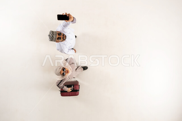 النظر الى الكاميرا بإيماءات السرور، الخدمة المتطورة لبرنامج السياحة والسفر, الاستعداد لرحلة ترفيهية ممتعة, بورتريه علوي عامودي لشاب عربي خليجي عماني يرتدي الدشداشة و الكمه يقوم بعرض الهاتف المحمول بشاشة سوداء فارغة، سيدة محجبة تمسك حقيبة السفر، خلفية بيضاء