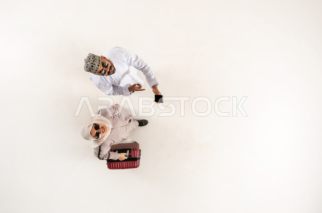 النظر الى الكاميرا بإيماءات السرور، الخدمة المتطورة لبرنامج السياحة والسفر, الاستعداد لرحلة ترفيهية ممتعة, بورتريه علوي عامودي لشاب عربي خليجي عماني يرتدي الدشداشة و الكمه يشير بإصبع السبابة الى الهاتف المحمول يقف مع زوجته، سيدة محجبة تمسك حقيبة السفر، خلفية بيضاء