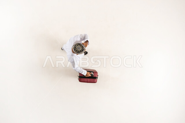 استخدام التقنية الحديثة والتطبيق السياحي, الخدمة المتطورة لبرنامج السياحة والسفر, بورتريه علوي عامودي لشاب عربي خليجي عماني يرتدي الدشداشة و الكمه يمسك حقيبة السفر والهاتف المحمول يقوم بإجراء مكالمه صوتية, الاستعداد لرحلة ترفيهية ممتعة, خلفية بيضاء