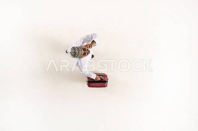 Advanced service for tourism and travel program, use of modern technology and tourism application, vertical upper portrait of a young Arab Gulf Omani man wearing a dishdasha and a sleeve holding a travel bag and a mobile phone making a voice call, preparing for a fun recreational trip, white background