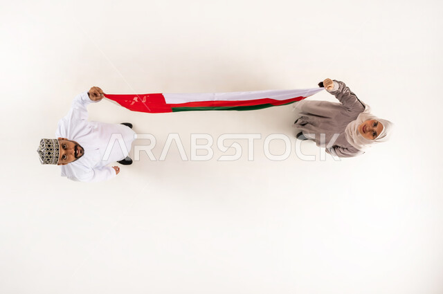 إظهار الولاء و الاخلاص والانتماء للبلاد، الاحتفال باليوم الوطني العماني 18 نوفمبر، أعياد ومناسبات وطنية، بورتريه علوي عامودي لرجل عربي خليجي عماني يرتدي الدشداشة والكمه يقف مع زوجته يحملان علم عمان ينظران الى الكاميرا بإيماءات السرور، خلفية بيضاء