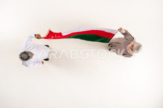 أعياد ومناسبات وطنية، إظهار الولاء و الاخلاص والانتماء للبلاد، الاحتفال باليوم الوطني العماني 18 نوفمبر، بورتريه علوي عامودي لرجل عربي خليجي عماني يرتدي الدشداشة والكمه يقف مع زوجته يحملان علم عمان ينظران الى بعضهما بإيماءات السرور، خلفية بيضاء