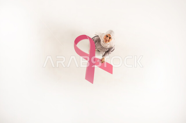 تقديم المساعدة والدعم من خلال الكشف المبكر عن سرطان الثدي, بورتريه علوي عامودي لشابة عربية خليجية عمانية مبتسمة محجبة ترتدي العباءة تمسك بيدها شعار  الحملة العالمية الوردي لتحدي الأمراض الخبيثة، سيدة سعودية تنظر الى الكاميرا بإيماءات السرور, خلفية بيضاء