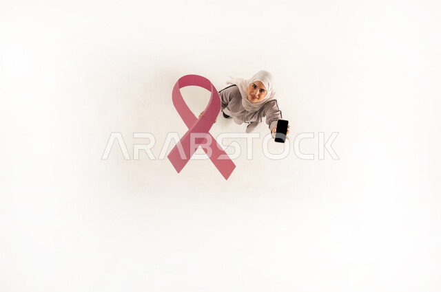 التوعيه والتثقيف والتحفيز، اليوم العالمي للكشف المبكر عن سرطان الثدي, بورتريه عامودي علوي لسيدة عربيه خليجيه عمانية ترتدي الحجاب والعباءة تمسك بيدها شعار اليوم العالمي لسرطان الثدي، امراة سعودية تقوم بعرض هاتف محمول بشاشة سوادء، اهميه اجراء الفحص المبكر، خلفية بيضاء