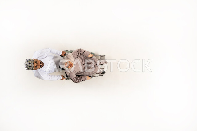 الاهتمام بالمعاقين وتقديم الدعم النفسي لهم, حاجة ذوي الهمم إلى الرعاية الصحية الخاصة, بورتريه عامودي علوي لزوج عربي خليجي عماني يرتدي الدشداشة والكمه يقوم بجر زوجته المقعدة وينظران الى الاعلى، ايماءات الحنان و العطف والشعور بالمحبة، خلفيه بيضاء