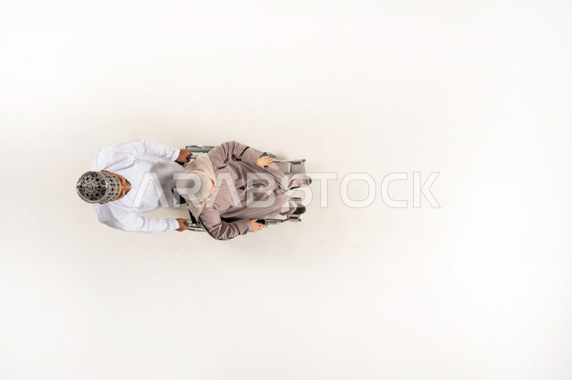 ايماءات الحنان و العطف والشعور بالمحبة، الاهتمام بالمعاقين وتقديم الدعم النفسي لهم, حاجة ذوي الهمم إلى الرعاية الصحية الخاصة, بورتريه عامودي علوي لزوج عربي خليجي عماني يرتدي الدشداشة والكمه يقوم بجر زوجته المقعدة، خلفيه بيضاء