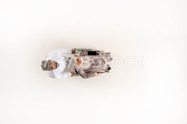 الاهتمام بالمعاقين وتقديم الدعم النفسي لهم, حاجة ذوي الهمم إلى الرعاية الصحية الخاصة, بورتريه عامودي علوي لزوج عربي خليجي عماني يرتدي الدشداشة والكمه يقوم بجر زوجته المقعدة وينظران الى الاعلى، ايماءات الحنان و العطف والشعور بالمحبة، خلفيه بيضاء