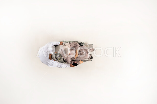 Mobile browsing, care for the disabled and providing them with psychological support, people with disabilities need special health care, vertical upper portrait of an Arab Gulf Omani husband wearing a dishdasha and a cloak pulling his disabled wife, gestures of tenderness and care, compassion and a sense of interest, white background