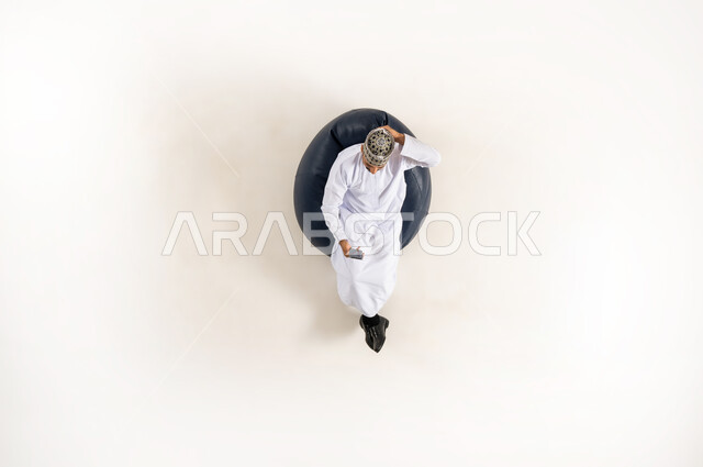 التسويق الإلكتروني وعرض خدمات وتطبيقات الجوال، إستخدام التكنولوجيا والتقنية الحديثة، بورتريه عامودي علوي لرجل عربي خليجي عماني يرتدي الدشداشة والكمه يجلس على كنبه مريحه ويتصفح الهاتف المحمول، خلفية بيضاء