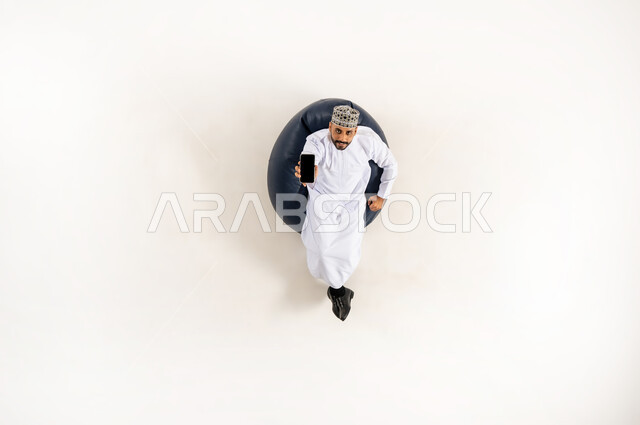 E-marketing and displaying mobile services and applications, using modern technology and techniques, vertical portrait of an Arab Gulf Omani man wearing a dishdasha and a turban sitting on a comfortable sofa raising his mobile phone up with a blank black screen, white background