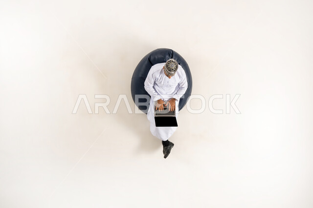 الاستفادة من التكنولوجيا والتقنيات المتطورة، بورتريه عامودي علوي لرجل عربي خليجي عماني يرتدي الدشداشة والكمه، الجلوس و العمل على الحاسوب المحمول، قضاء الوقت مع الذات، انجاز المهام عن بعد، خلفية بيضاء