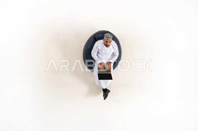 انجاز المهام عن بعد، الاستفادة من التكنولوجيا والتقنيات المتطورة، بورتريه عامودي علوي لرجل عربي خليجي عماني يرتدي الدشداشة والكمه، الجلوس و العمل على الحاسوب المحمول، قضاء الوقت مع الذات، خلفية بيضاء