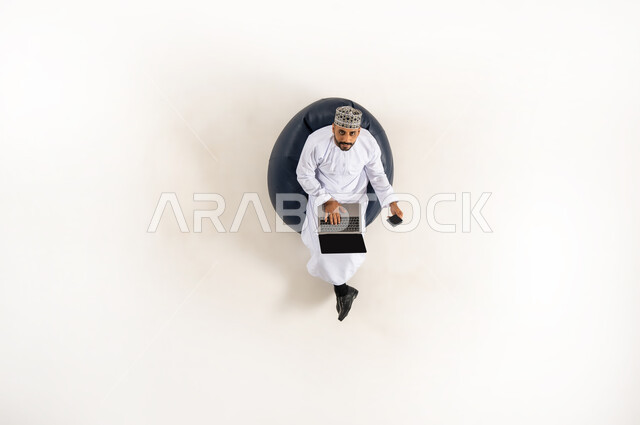سهولة السحب عبر الحساب البنكي, استخدام التكنولوجيا المتطورة والتقنيات الحديثة, بورتريه عامودي علوي لرجل عربي خليجي عماني يرتدي الدشداشة والكمه ينظر الى الاعلى ويقوم بالدفع الالكتروني بواسطة الحاسوب المحمول, استخدام بطاقة الصراف,  خلفية بيضاء