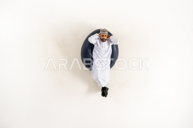 متعة قضاء الوقت مع الذات، الاسترخاء والتأمل في اوقات الفراغ،  بورتريه عامودي علوي لرجل عربي خليجي عماني يرتدي الدشداشة والكمه مستلقي على كنبه مريحه ويضع يديه خلف رأسه، ايماءات تدل على الراحة والسرور، خلفية بيضاء
